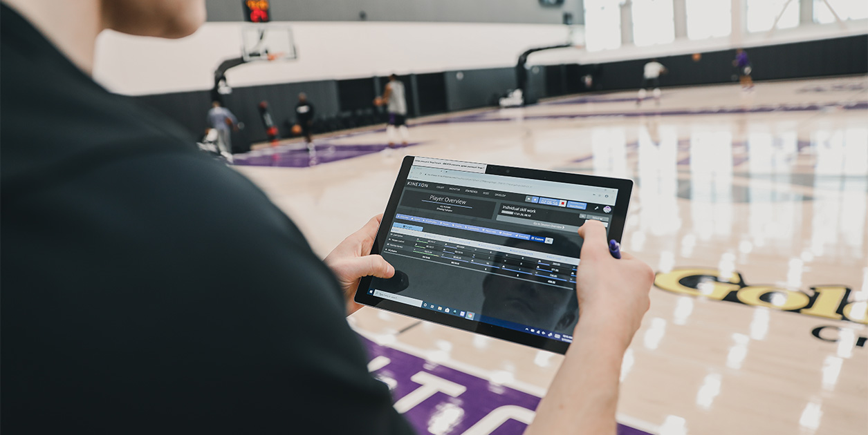 A man holding a tablet checks sporting performance data while he watches basketball training.