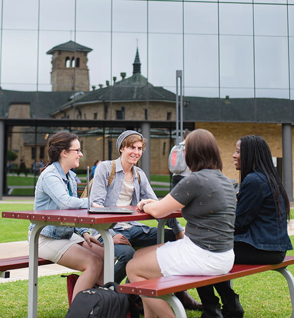 Students sitting around an outdoor table with the reflection of a campus building behind them