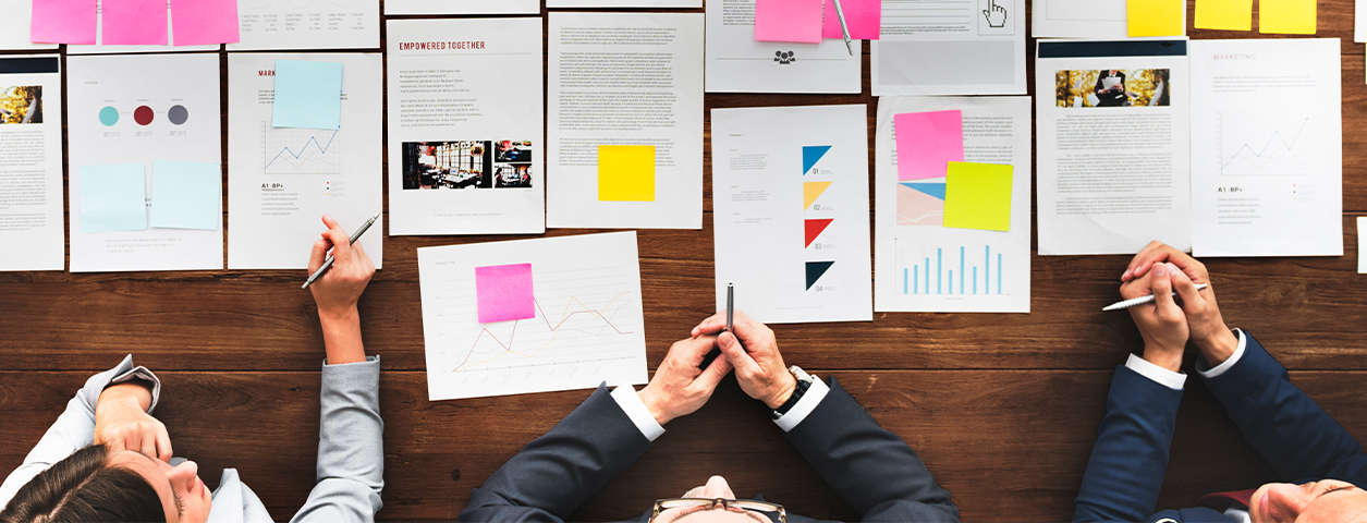Three professionals sit at one edge of a wooden table, looking over notes and presentations