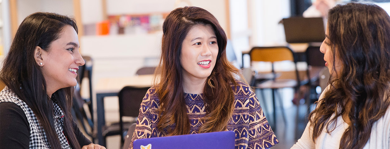 Three women sit at a desk talking.