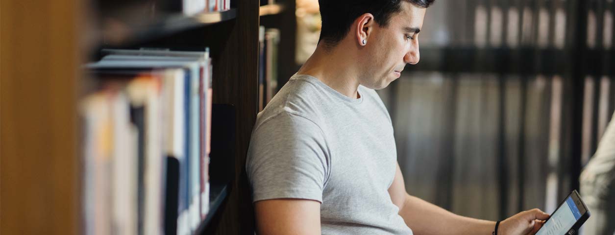 Man in grey t-shirt using a laptop whilst sitting in a library.
