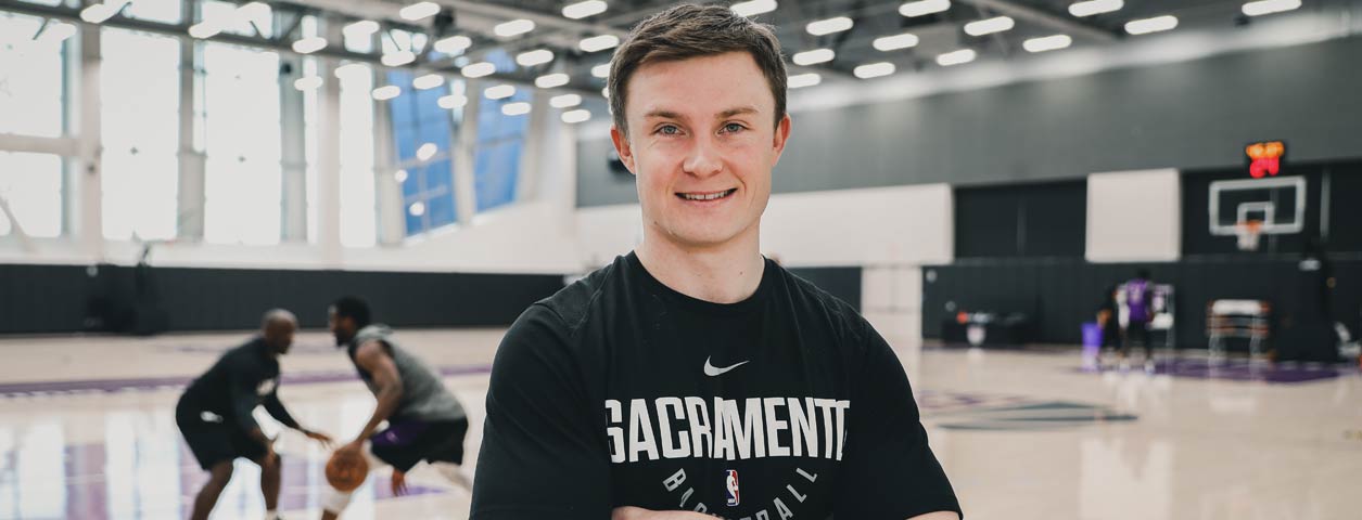 Smiling basketball player with arms folded, standing infront of the camera with a basketball game being played in the background inside a basketball stadium.