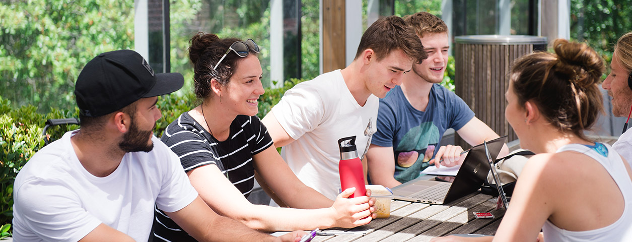 Several students gather on an outdoor table with laptops and books, talking