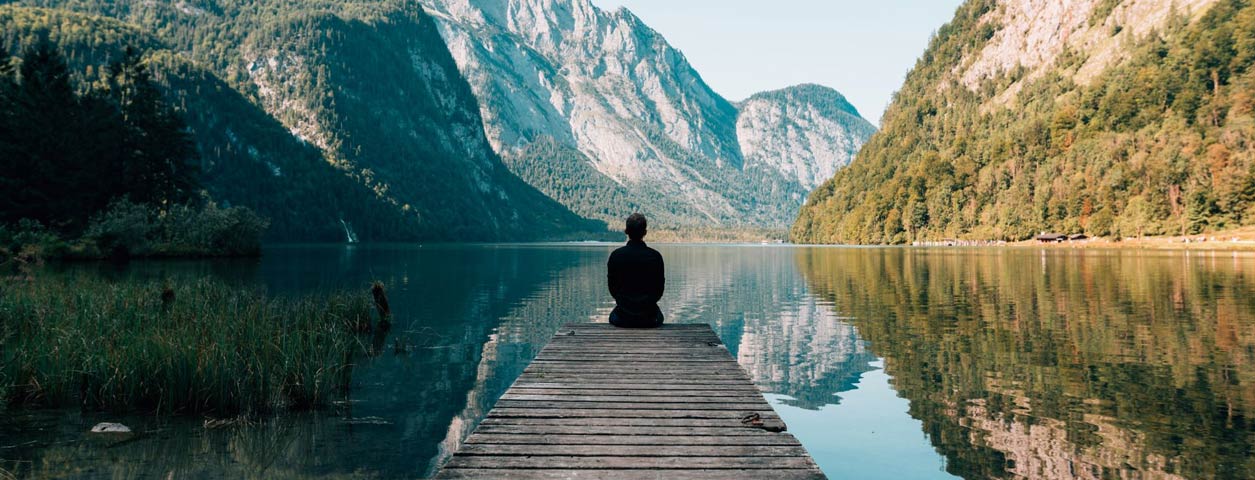 Person sitting on a pier of a small lake surrounded by mountains.