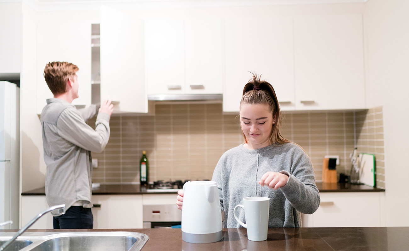 A managed residence kitchen. It is clean and new.  In the foreground, a student is making herself a cup of tea while behind her another student reaches into a cupboard.