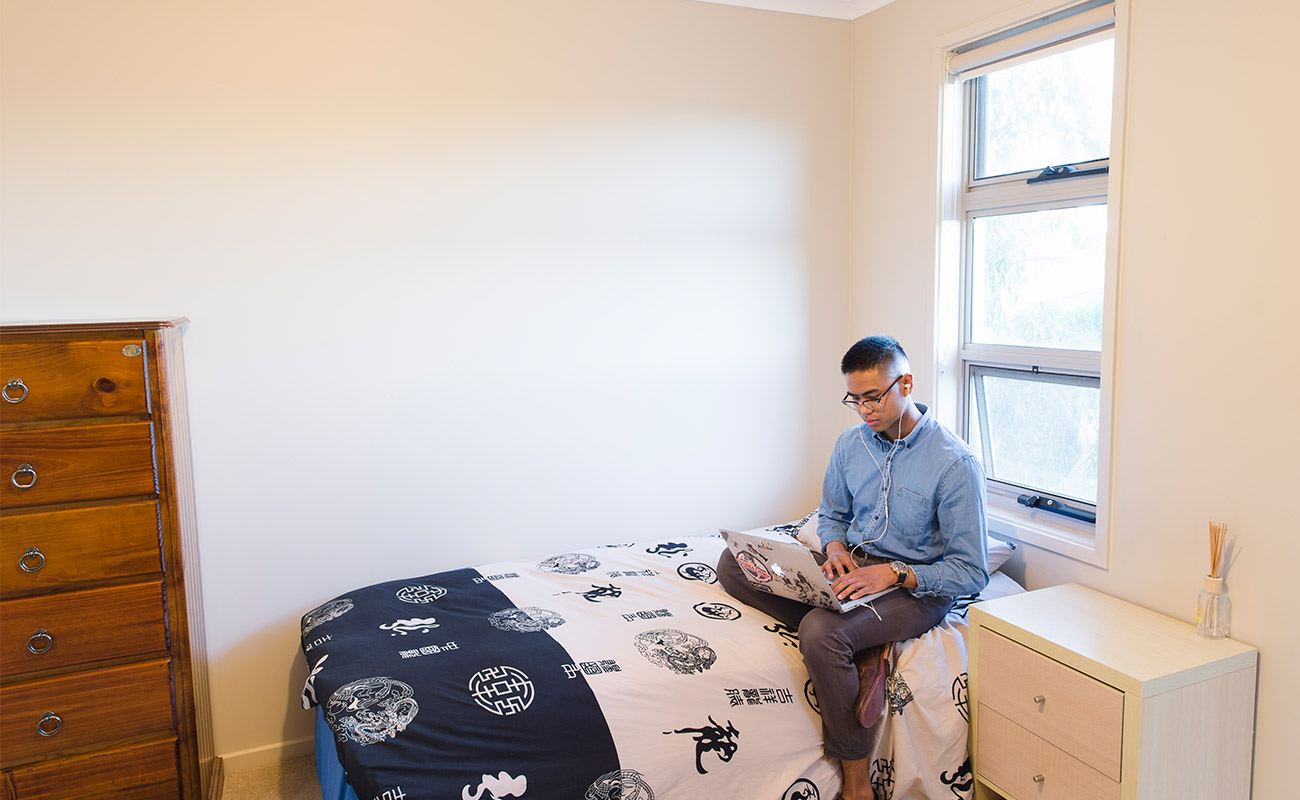 A student sits on his bed in the managed residence, listening to something on his laptop. There is a window at the head of the bed and a small drawer beside the single bed.