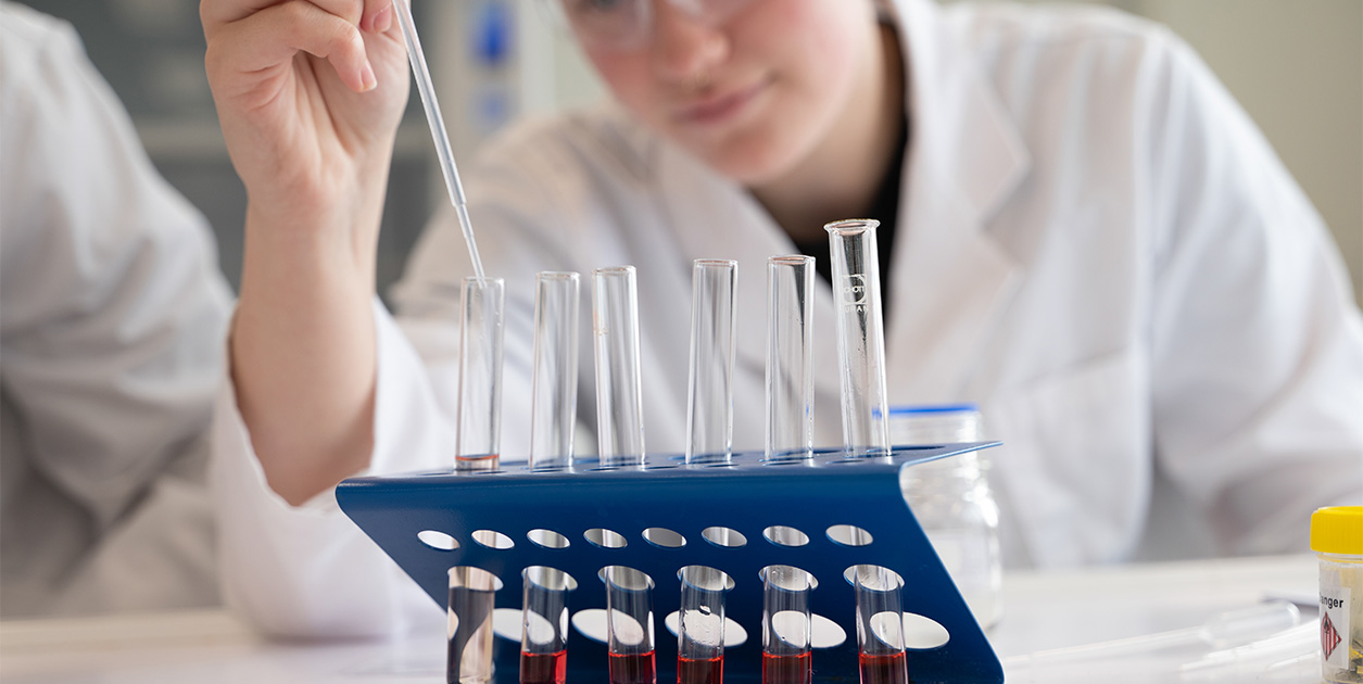 A student carefully puts liquid into a test tube standing in a rack of several test tubes.