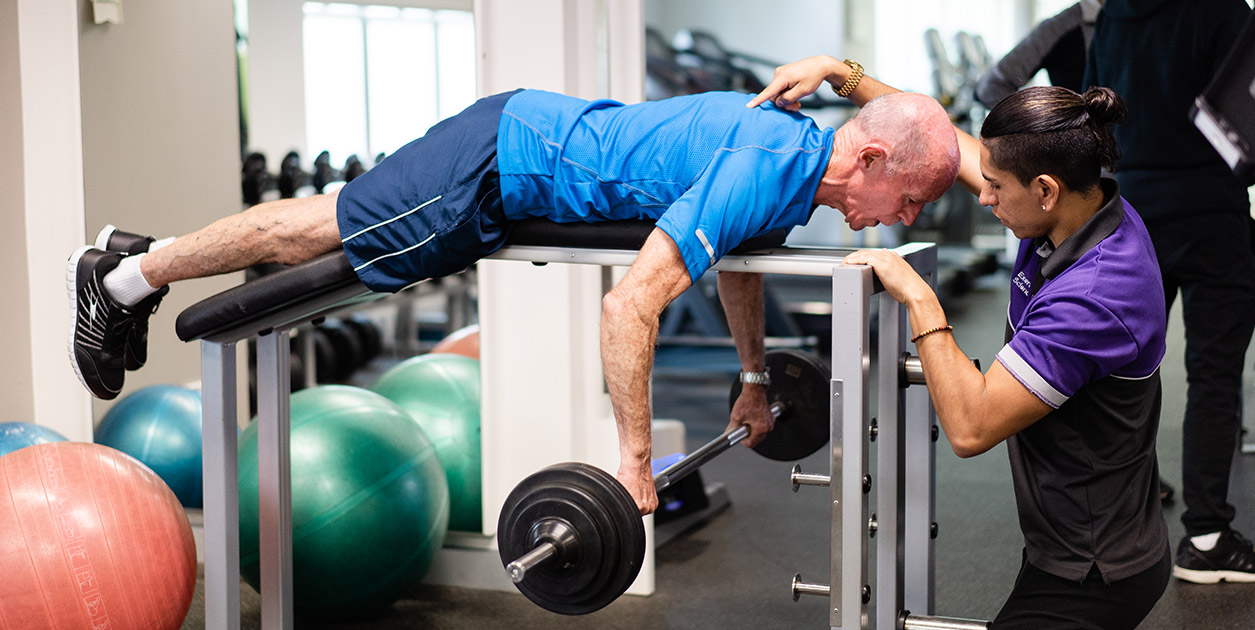 Clinical exercise student assists an elderly patient with a barbell