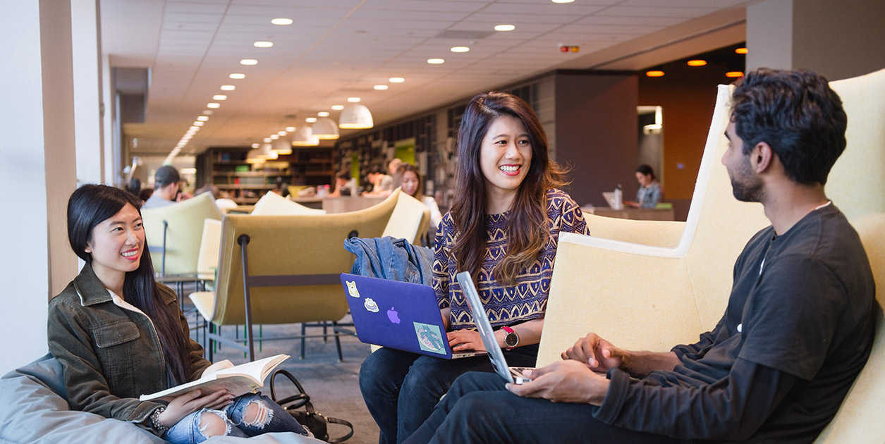 Three students sit in a study area with laptops and books