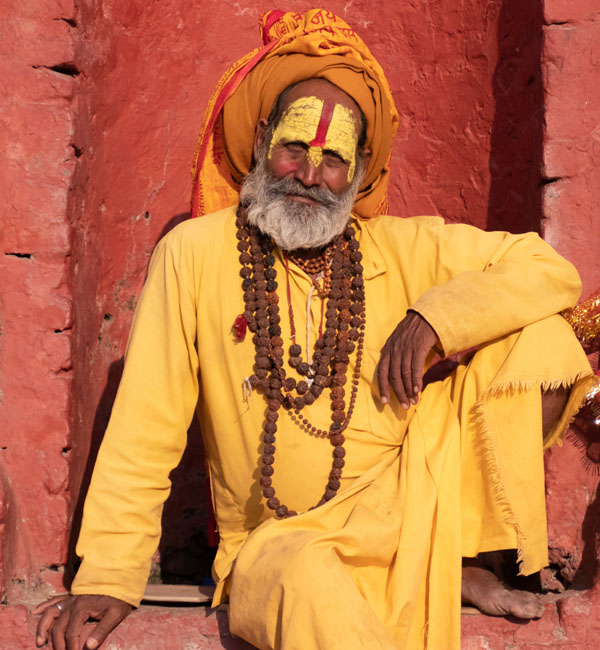 Seated Pujar wearing yellow clothing and face paint with neck beads and a dark orange turban