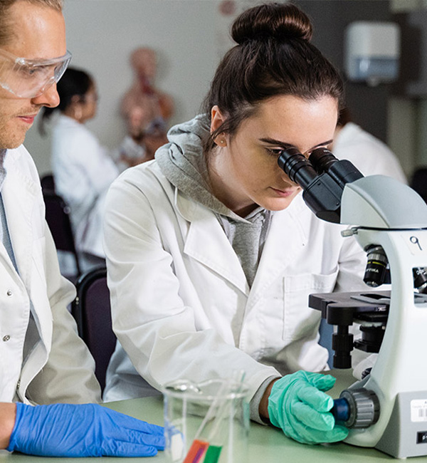 A student in a white coat and green gloves looks through a microscope