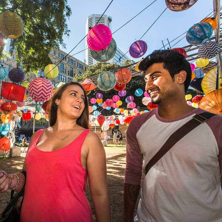Two people walking through an outdoor installation filled with paper lanterns suspended above them.